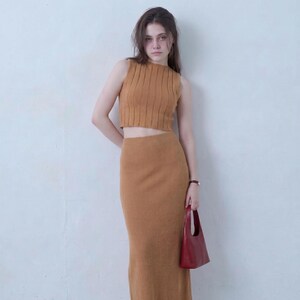 Puede incluir: Un conjunto de dos piezas color amarillo mostaza compuesto por un top corto sin mangas acanalado y una falda larga a juego de cintura alta, acompañado de un pequeño bolso de mano rojo oscuro. La prenda presenta una textura acanalada vertical.