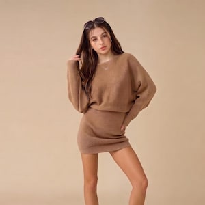Puede incluir: Un vestido de punto color canela de manga larga con textura acanalada, cuello redondo y silueta ajustada. La prenda se combina con gafas de sol de montura oscura colocadas sobre la cabeza, creando un aspecto monocromático y uniforme.