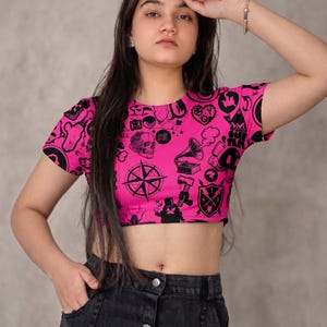 Puede incluir: Un top corto rosa intenso con un estampado gráfico negro con varios símbolos e iconos. El top tiene mangas cortas y cuello redondo. La modelo también lleva una falda vaquera negra.
