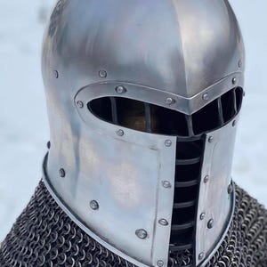 May include: A polished silver medieval helmet with a rounded top and a visor featuring vertical slits for vision. The helmet is riveted and sits atop a chainmail coif, offering historical protection.