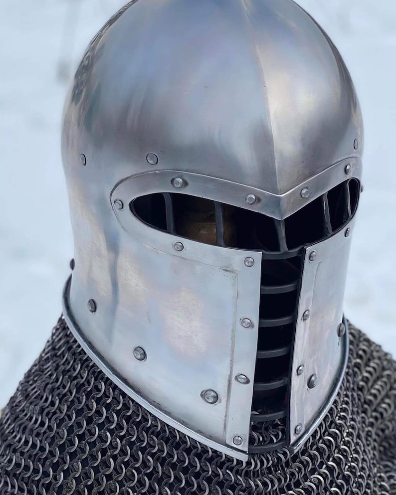 May include: A polished silver medieval helmet with a rounded top and a visor featuring vertical slits for vision. The helmet is riveted and sits atop a chainmail coif, offering historical protection.