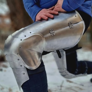 Könnte beinhalten: Eine polierte, silberfarbene Metallknie- und Unterschenkelpanzerung. Die Panzerung ist genietet und scharniert, mit einem gebogenen Design zum Schutz des Kniegelenks. Der untere Beinabschnitt erstreckt sich bis zum Knöchel.