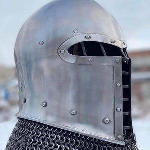 May include: A polished, silver-colored medieval helmet with riveted construction. The helmet features a rounded top, a face guard with vertical bars, and a chainmail neck protector. The design is reminiscent of historical armor.