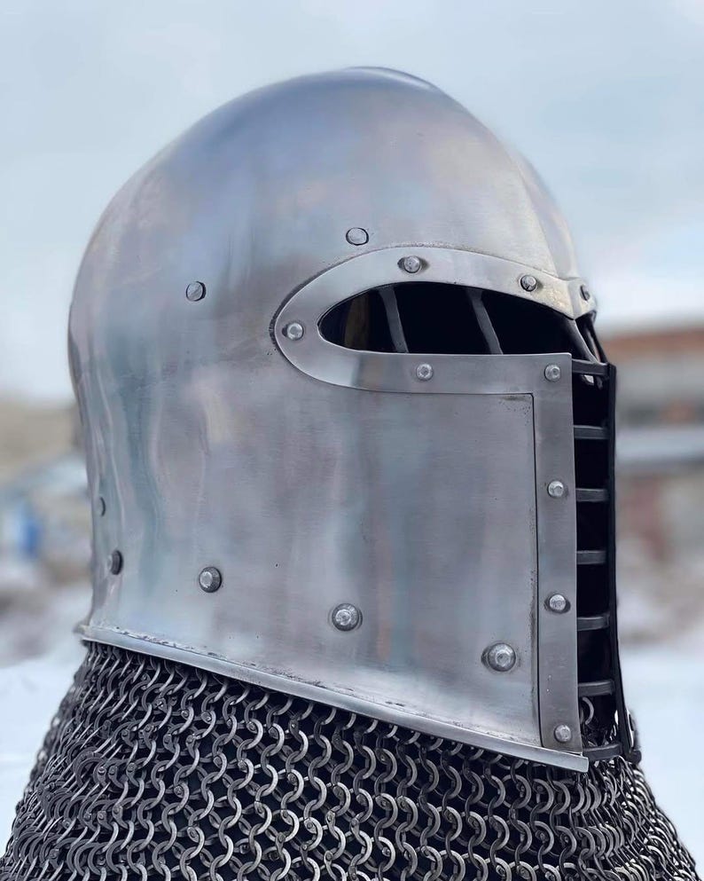 May include: A polished, silver-colored medieval helmet with riveted construction. The helmet features a rounded top, a face guard with vertical bars, and a chainmail neck protector. The design is reminiscent of historical armor.