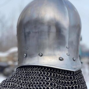 May include: A polished, silver-colored medieval helmet with a rounded top and riveted brim. The helmet sits atop a chainmail coif, creating a historical aesthetic. The image is taken from a slightly low angle, emphasizing the helmet's shape.