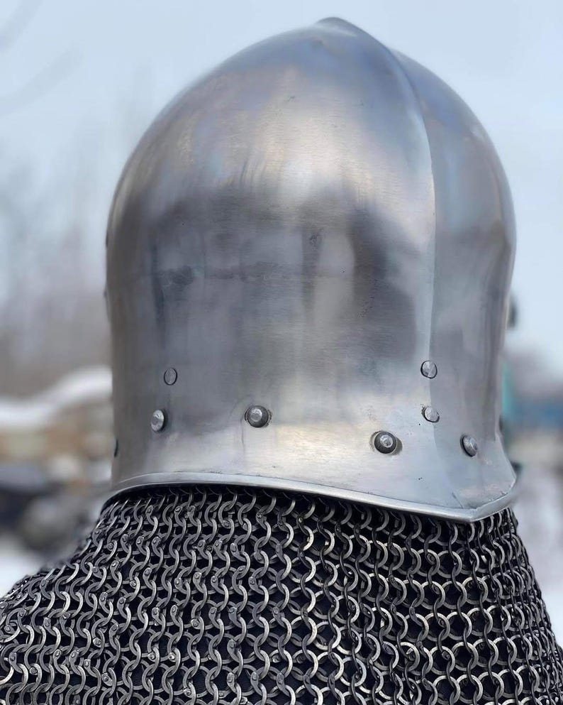 May include: A polished, silver-colored medieval helmet with a rounded top and riveted brim. The helmet sits atop a chainmail coif, creating a historical aesthetic. The image is taken from a slightly low angle, emphasizing the helmet's shape.