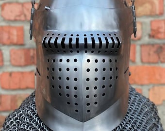 May include: A silver-coloured medieval helmet with a visor featuring horizontal slits and circular ventilation holes. The helmet sits above a chainmail coif, with a brick wall in the background. This historical armour piece is likely for costume or display.