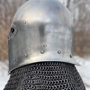 May include: A polished, silver-colored medieval helmet with a rounded top and riveted details. The helmet features a visor and is paired with a chainmail coif. The background is blurred, suggesting an outdoor setting.