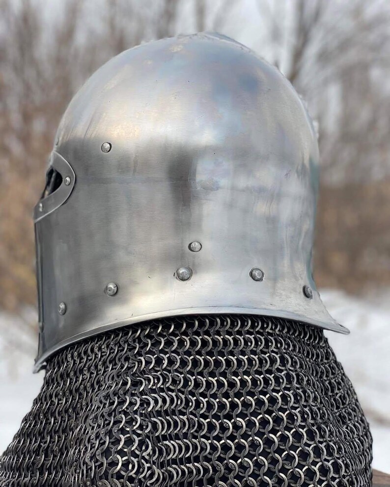 May include: A polished, silver-colored medieval helmet with a rounded top and riveted details. The helmet features a visor and is paired with a chainmail coif. The background is blurred, suggesting an outdoor setting.