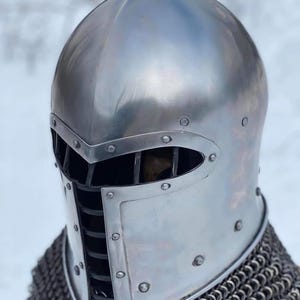 May include: A polished silver medieval helmet with a visor and riveted construction. The helmet features a rounded top and a protective face guard with vertical bars. Chainmail is visible at the base of the helmet.