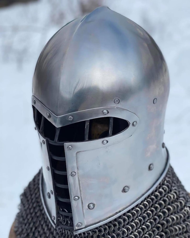 May include: A polished silver medieval helmet with a visor and riveted construction. The helmet features a rounded top and a protective face guard with vertical bars. Chainmail is visible at the base of the helmet.