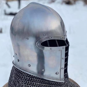 May include: A polished steel medieval helmet with riveted construction. The helmet features a visor with eye slits and a neck guard made of chainmail. The background is a snowy outdoor scene.