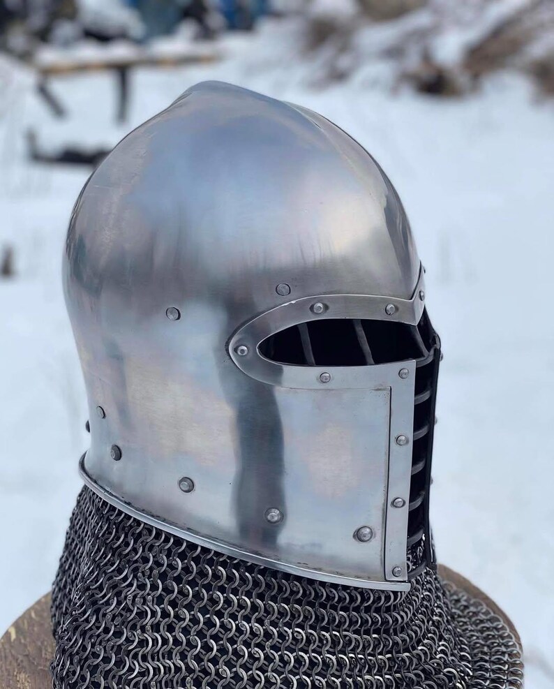 May include: A polished steel medieval helmet with riveted construction. The helmet features a visor with eye slits and a neck guard made of chainmail. The background is a snowy outdoor scene.