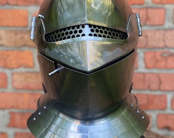 May include: A polished steel knight's helmet with a visor and neck guard. The helmet features a series of small holes for ventilation and is designed to protect the head and neck. The helmet is displayed on a wooden surface against a brick wall.