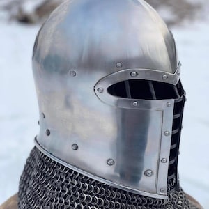 May include: A polished, silver-colored medieval helmet with riveted construction. The helmet features an eye slit with vertical bars and is paired with a chainmail coif. The design is reminiscent of historical armor.