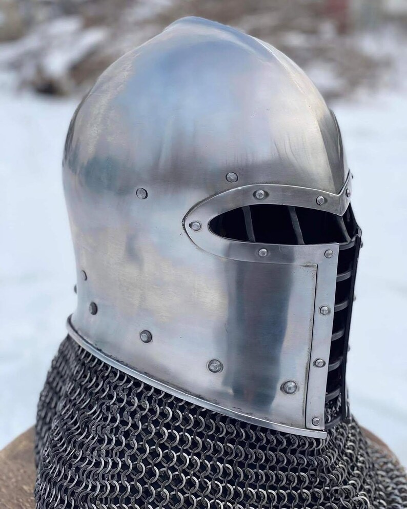 May include: A polished, silver-colored medieval helmet with riveted construction. The helmet features an eye slit with vertical bars and is paired with a chainmail coif. The design is reminiscent of historical armor.
