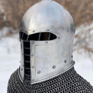 May include: A polished silver medieval helmet with a riveted construction and a barred visor. The helmet is worn over a chainmail coif, offering protection for historical reenactment or display.