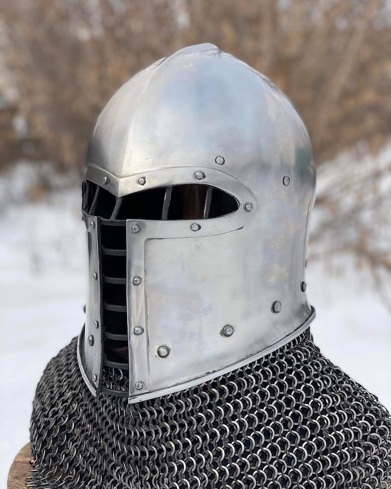 May include: A polished silver medieval helmet with a riveted construction and a barred visor. The helmet is worn over a chainmail coif, offering protection for historical reenactment or display.