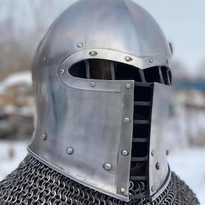 May include: A silver-colored medieval helmet with riveted construction and a visor featuring horizontal slits for vision. The helmet is secured with rivets and is worn over a chainmail coif.