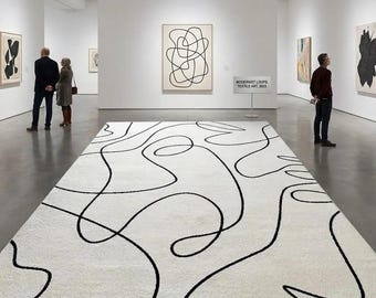 Puede incluir: Una alfombra blanca con un diseño de líneas negras abstractas domina el primer plano de una galería de arte. El diseño de la alfombra refleja las obras de arte en las paredes. La galería presenta pinturas abstractas enmarcadas y un letrero que dice "Modernist Loops, Textile Art 2023."