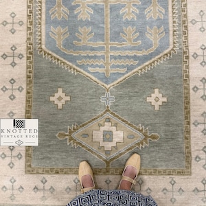 Op de afbeelding: Een rechthoekig vloerkleed met een saliegroen veld en een centraal lichtblauw zeshoekig medaillon met een beige boommotief. De rand heeft een geometrisch patroon en het omliggende crèmekleurige veld bevat kleine kruisvormige symbolen.