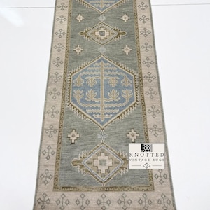 May include: A long rectangular runner rug featuring a sage green central field with two large blue hexagonal medallions containing tree motifs. The border is a light beige with repeating geometric patterns and a white fringe at both ends.