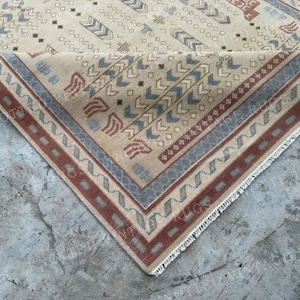 May include: A beige area rug featuring a geometric pattern in shades of blue and terracotta red. The rug has a folded corner revealing a white fringe edge and a border with a repeating zigzag design. The text KNOTTED VINTAGE RUGS is visible.