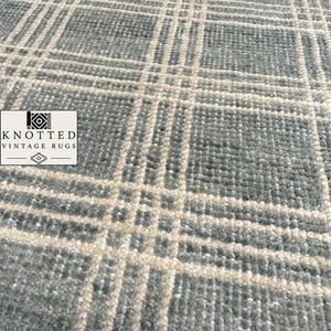 Peut inclure: Un tapis présentant un motif à carreaux bleu et blanc avec une surface à poils noués texturés. Un logo dans le coin affiche le texte KNOTTED VINTAGE RUGS avec une icône géométrique au-dessus et un petit symbole décoratif sous le texte.