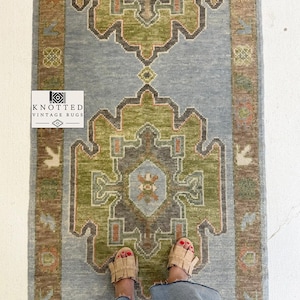 May include: A rectangular vintage rug with a central design in shades of green, brown, and orange, set against a light blue background. The rug has a border in shades of brown and blue. The rug has the text "KNOTTED VINTAGE RUGS".
