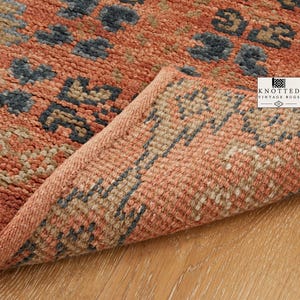 May include: A woven rug featuring a terracotta orange base with a repeating geometric pattern in dark charcoal and beige. The corner of the rug is folded over, revealing the textured underside and the logo text KNOTTED VINTAGE RUGS.