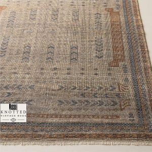 Puede incluir: Una alfombra rectangular con base beige y un patrón repetitivo de formas geométricas azules y color óxido, incluyendo flechas y diamantes. La alfombra tiene un borde con flecos y un logotipo en la esquina que dice KNOTTED VINTAGE RUGS.