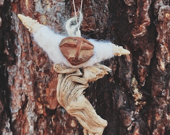 May include: A handmade wooden ornament with a carved face, white felt wings, and a driftwood body. The ornament hangs from a white string against a textured brown background, likely a tree trunk. The rustic design suggests a nature-inspired theme.