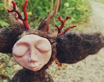 May include: A handcrafted figurine featuring a pale, smooth, sculpted face with closed eyes and a subtle smile. The head is covered in dark brown, textured wool fibers, accented by two bright red, branch-like structures resembling antlers.