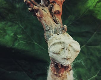 May include: A sculpture featuring a rough, brown wooden branch base topped with a pale, off-white molded face. The face has closed eyes and a defined nose. Below the face, a textured, light beige fibrous material covers the lower section.
