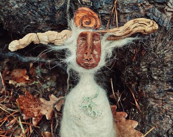 May include: A handcrafted forest spirit doll with a carved face, wooden arms, and a white wool body. The doll is adorned with a spiral headpiece and a sprig of green lichen. It rests among fallen leaves and pine needles.