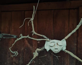 Puede incluir: Un colgante de pared hecho a mano y caprichoso que presenta una escultura de rostro blanco y sereno unida a una disposición en forma de cruz de ramitas naturales. La escultura está suspendida por una fina cuerda blanca sobre un fondo de madera rústica.