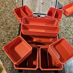 May include: An open black and red toolbox with several removable orange storage bins. The bins are various sizes and shapes, designed to organise tools or small items. The toolbox is on a light-coloured surface.