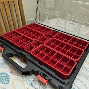 Peut inclure: Une mallette à outils en plastique noir et transparent avec des compartiments de rangement rouges. L'étui est ouvert, révélant plusieurs plateaux rouges avec des compartiments de différentes tailles. L'étui a une poignée rouge et des loquets argentés.