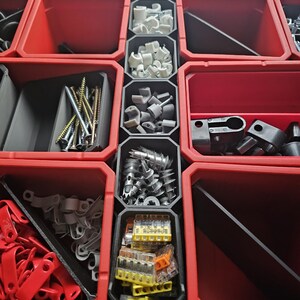 Può includere: Una vista ravvicinata di un contenitore di stoccaggio organizzato pieno di vari articoli hardware. Il contenitore presenta scomparti rossi e grigi che contengono viti, connettori per cavi e altre piccole parti. Gli articoli sono ben organizzati.