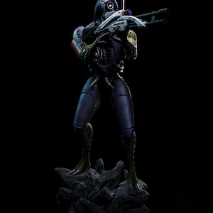 May include: A detailed figurine of a science fiction character in a dark blue and black suit, holding a futuristic rifle. The figure stands on a rocky base against a black background. The rifle is white and gray with red and blue accents.