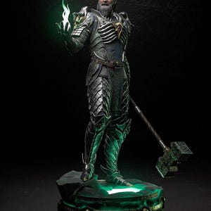Puede incluir: Estatua detallada de un guerrero con armadura plateada y verde, sosteniendo una llama verde brillante. La figura tiene una larga barba blanca y ojos verdes. Un gran martillo descansa junto a la figura. La estatua está sobre una base verde iluminada.