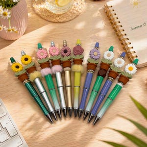 Könnte beinhalten: Eine Sammlung dekorativer Stifte mit blütenförmigen Aufsätzen. Die Stifte haben verschiedene Farben, darunter Grün, Silber, Gold und Lila. Jeder Stift hat ein kleines Topfdesign und eine andere Blume, wie Sonnenblumen, Gänseblümchen und Rosen.