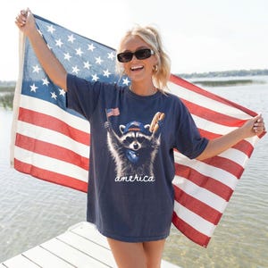 Pode incluir: Uma t-shirt cinzenta escura de manga curta com um gráfico de um guaxinim a usar óculos de sol, um boné de basebol e uma bandana. O guaxinim segura uma pequena bandeira americana e um cachorro-quente. A palavra america está escrita em branco.