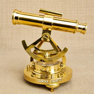 May include: A polished brass telescope with a compass base. The telescope has a cylindrical barrel and is mounted on a detailed brass stand. The base is engraved with "URVA CRAFT".