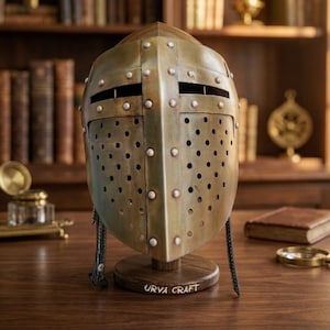 May include: A brass-toned metal knight helmet featuring a cross-shaped front plate with numerous circular ventilation holes and silver-colored rivets. The helmet is mounted on a circular wooden base with the text Urva Craft engraved on the front.