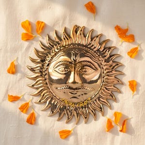 Peut inclure: Un accrochage mural doré en forme de soleil avec des traits de visage détaillés et des rayons rayonnants. Des pétales de fleurs orange sont dispersés autour du soleil sur une surface blanche. Le soleil a une moustache et une expression sereine.