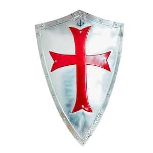 Peut inclure: Un bouclier argenté avec une croix rouge proéminente au centre. Le bouclier a une base pointue et un sommet incurvé. Le texte "URVA CRAFT" est visible le long du bord inférieur. Une ancre est visible en haut.