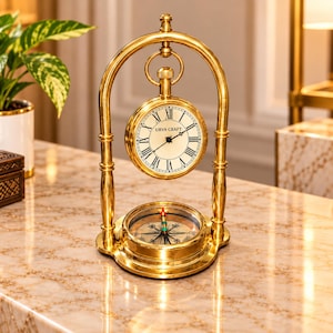 May include: A gold-toned metal desk clock and compass set featuring an arched frame. The clock has a white face with black Roman numerals and the text URVA CRAFT. A circular compass is positioned at the base of the arched stand.