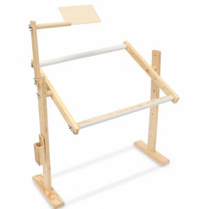 Puede incluir: Un soporte de bordado de madera con un marco rectangular y una pequeña estantería adjunta. El soporte tiene ajustes de altura y ángulo ajustables, con barras acolchadas blancas para sujetar la tela. El color de la madera natural le da un aspecto clásico.