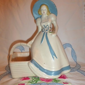 May include: A vintage ceramic planter in the shape of a woman wearing a blue hat and dress. The planter has a basket-shaped base.